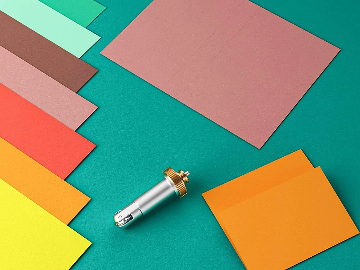 Papiers de couleur — jaune, orange, corail, menthe, rose poudré — disposés en éventail sur un fond vert, à côté de l’outil de rainage Wellkitch.